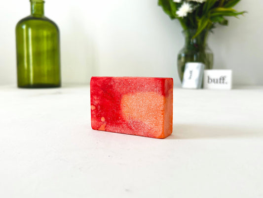 A red and orange rectangle bar soap, placed on a white surface. A large green vase and fresh flowers are placed behind the soap